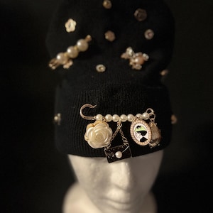 May include: A black beanie with a variety of decorative charms, including a pearl necklace, a rose, a purse, and a portrait. The charms are attached to a safety pin.