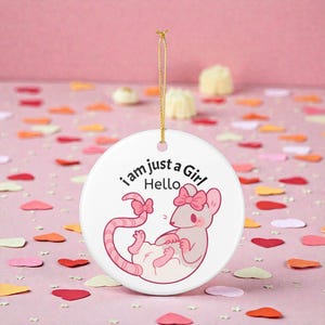 Könnte beinhalten: Weißes Keramik-Ornament mit einer Cartoon-Illustration einer rosa Maus mit Schleife. Der Text "i am just a Girl Hello" ist aufgedruckt. Ein goldener Faden dient zum Aufhängen. Der Hintergrund ist rosa mit Konfetti.