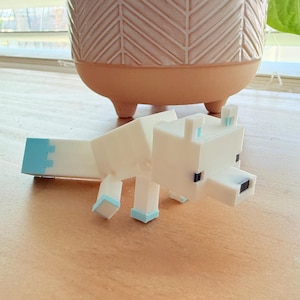 May include: A white, geometric-style wolf figurine with light blue accents on its ears, tail, and feet. The figurine is positioned on a wooden surface, with a decorative plant pot in the background.