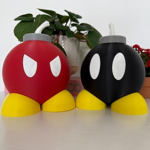 Pode incluir: Duas figuras de bomba em estilo cartoon, uma vermelha e outra preta, com olhos brancos e pés amarelos. A bomba vermelha tem uma expressão zangada. Ambas têm tampas cinzentas e pavios brancos. As figuras estão numa superfície branca.