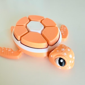 以下が含まれることがあります： オレンジと白の3Dプリントされた亀の玩具。セグメント化されたシェルと頭とひれに白い水玉模様があります。