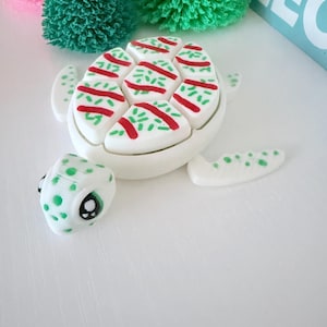 May include: A decorative white turtle figurine with a shell featuring red stripes and green sprinkles. The turtle's head and flippers are also white with green spots. The figurine is on a white surface, with green and pink decorative trees in the background.