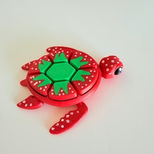以下が含まれることがあります： 白のドット模様がついた赤と緑の3Dプリントされた亀の玩具。亀の甲羅は緑色の芯を持つイチゴの形をしています。