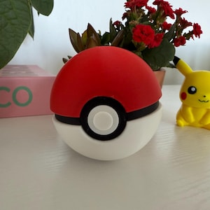 May include: A red and white Poké Ball with a black band and white button, next to a yellow Pikachu figurine. A pink box with the word "XICO" is visible in the background.