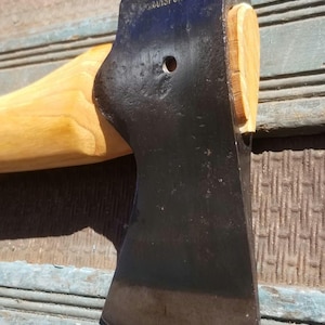 May include: A metal axe head with a dark, weathered finish and the text GRANSFORS stamped on the top edge. The axe head features a circular hole in the center and is attached to a light-colored wooden handle with a wooden wedge visible at the top.