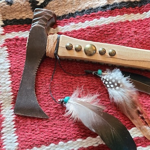 May include: A handcrafted tomahawk with a dark metal blade and a light wood handle adorned with brass studs. The handle is wrapped in leather. Decorative feathers with turquoise beads hang from the handle. The tomahawk rests on a red and white patterned textile.