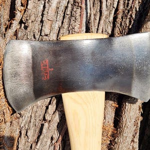 May include: A double-bit axe with a polished steel head and a wooden handle. The axe head has a red logo and the number "3" stamped on it. The axe is set against a textured tree bark background.