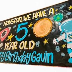 May include: A black banner with a space theme. It says "Houston, we have a 5 year old. Happy Birthday Gavin." There is a rocket, planets, stars, and an astronaut.