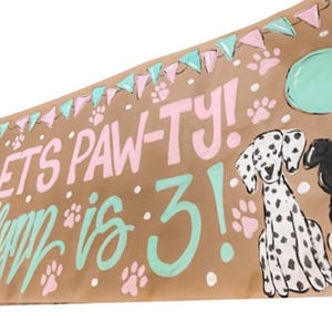 May include: A brown paper banner with the text "Let's Paw-ty! Happy is 3!" in pink and green paint. The banner also features illustrations of three dogs, a Dalmatian, a black and white border collie, and a brown and white dog wearing a party hat. There are also painted images of balloons and paw prints.