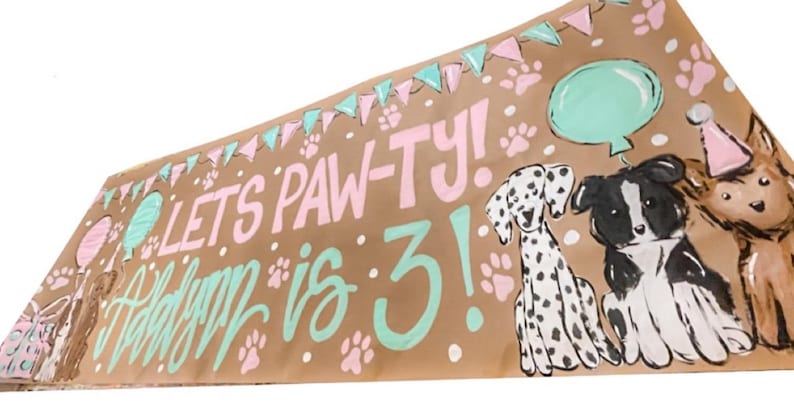 May include: A brown paper banner with the text "Let's Paw-ty! Happy is 3!" in pink and green paint. The banner also features illustrations of three dogs, a Dalmatian, a black and white border collie, and a brown and white dog wearing a party hat. There are also painted images of balloons and paw prints.