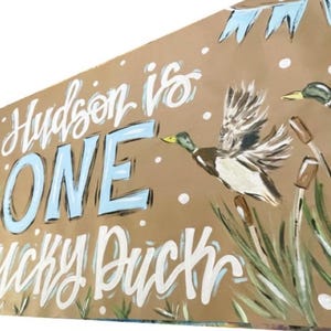 May include: A brown paper banner with the text "Hudson is ONE: Lucky Duck" in white and blue paint. The banner features two ducks in flight, painted in shades of brown, white, and blue, against a backdrop of green reeds.