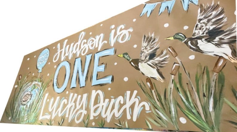 May include: A brown paper banner with the text "Hudson is ONE: Lucky Duck" in white and blue paint. The banner features two ducks in flight, painted in shades of brown, white, and blue, against a backdrop of green reeds.