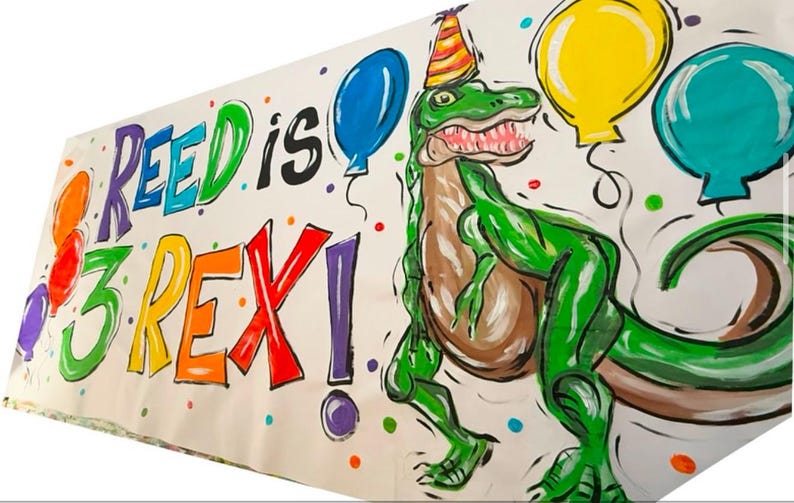 May include: A colorful banner with the text "Reed is 3 Rex!" in rainbow colors. A green dinosaur with a party hat is drawn on the banner with colorful balloons.