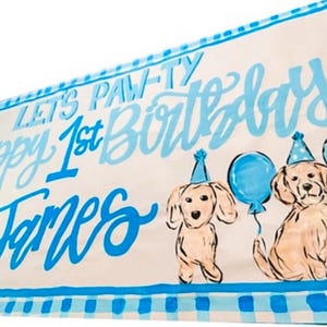 May include: A blue and white banner with the text "Let's Paw-ty Happy 1st Birthday James" and two cartoon dogs with blue balloons.
