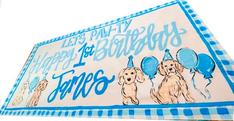 May include: A blue and white banner with the text "Let's Paw-ty Happy 1st Birthday James" and two cartoon dogs with blue balloons.