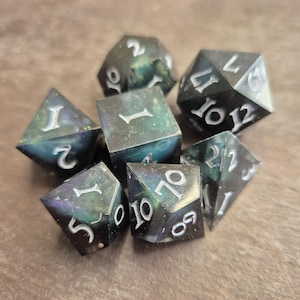 Könnte beinhalten: Ein Satz von sieben polyedrischen Würfeln für Tabletop-Spiele. Die Würfel sind dunkel, irisierend und mit weißen Zahlen versehen. Enthält einen W4, W6, W8, W10, W12, W20 und einen Prozentwürfel.