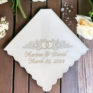 May include: A white handkerchief with scalloped edges featuring gold embroidery of two interlocking rings surrounded by floral flourishes. The text Marissa & David and the date March 23, 2024, are embroidered in gold script below the rings.
