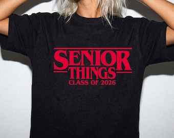 May include: Black t-shirt featuring the red text "SENIOR THINGS" with "CLASS OF 2026" underneath. The t-shirt has a crew neck design.