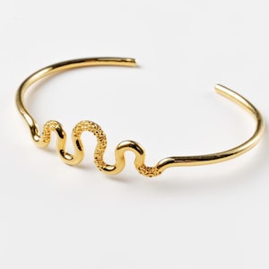 May include: A gold-coloured cuff bracelet with a wavy, textured design. The bracelet has an open design and a polished finish. The textured portion of the bracelet resembles a snake. The jewellery is displayed on a white background.
