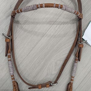 Può includere: Briglia per cavallo in pelle marrone con accenti intrecciati bianchi e grigi. La briglia presenta una capezzina, paraorecchie e un sottogola. Le redini sono attaccate con hardware in metallo. La briglia ha frange decorative.
