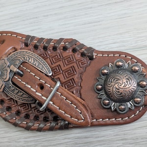 Può includere: Primo piano di un cinturino per speroni in pelle marrone con dettagli metallici decorativi. Il cinturino presenta una fibbia floreale, goffratura geometrica e una concho circolare ornata. Cuciture bianche e dettagli in pelle intrecciata.