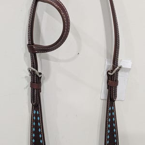 Può includere: Una briglia per cavalli in pelle marrone con cuciture bianche e dettagli a forma di diamante turchese. La briglia presenta fibbie color argento ed è progettata per l'uso equestre. La pelle sembra essere di alta qualità.