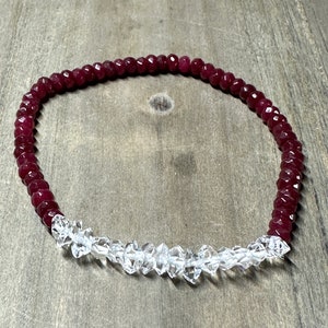 May include: A beaded bracelet featuring alternating sections of deep red ruby and clear quartz gemstones. The ruby beads are small and faceted, while the quartz beads are larger and irregularly shaped. The bracelet is strung on a clear elastic cord.
