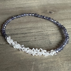Könnte beinhalten: Ein dehnbares Armband aus klaren Quarz- und Amethystperlen. Das Armband besteht aus facettierten Perlen und hat ein zweifarbiges Design.