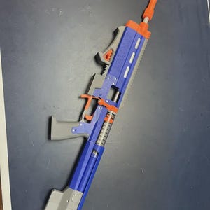 May include: A blue, orange, and gray toy gun with the word "DYNAIR" printed on the side. The toy gun has a long barrel and a magazine. The toy gun is made of plastic and appears to be a replica of a real firearm.