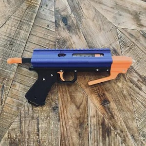 May include: A toy gun featuring a blue upper receiver, black grip, and orange details. The toy gun is made of plastic and rests on a wooden surface. The trigger and barrel tip are orange.