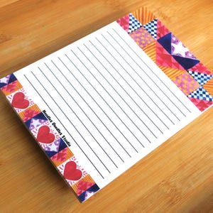 May include: A lined notepad with a decorative border featuring hearts, stars, and geometric patterns in red, pink, blue, and orange. The notepad is on a wooden surface, with the text "Noodles Doodles Studio" visible.