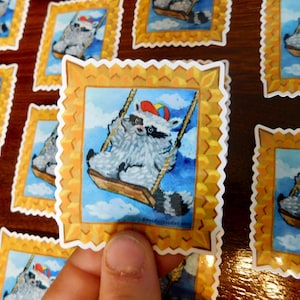 May include: A set of colorful stickers featuring a raccoon wearing a party hat and riding a swing. The stickers are printed on white paper with a gold frame and a scalloped edge.