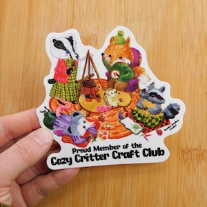May include: A die-cut sticker featuring a gathering of anthropomorphic animals. A badger, fox, raccoon, hedgehog, and opossum are depicted crafting and enjoying tea. The sticker reads "Proud Member of the Cozy Critter Craft Club."
