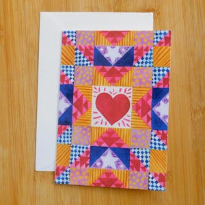 May include: A patterned greeting card with a quilt-inspired design. The card displays a central red heart, framed by geometric shapes in pink, blue, yellow, and orange. A white envelope accompanies the card, set against a wooden surface.
