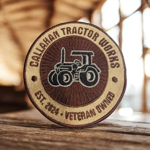 Może przedstawiać: Okrągła naszywka z napisem "Callahan Tractor Works" i ilustracją traktora. Naszywka jest brązowa i beżowa z napisem "Est. 2024 - Veteran Owned".