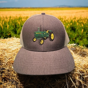 Può includere: Cappellino da camionista grigio con retro in rete e visiera curva. La parte anteriore del cappellino presenta un design ricamato di un trattore verde e giallo. Il cappellino è su una balla di fieno, con un campo sullo sfondo.