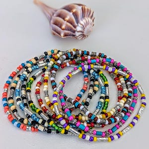 May include: A collection of colorful beaded necklaces with silver-toned metal accents. The beads are in various colors, including red, blue, yellow, and white. A brown and tan seashell is in the background.