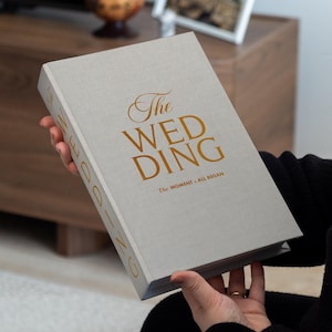 Op de afbeelding: Een lichtgrijs trouwalbum met gouden letters met de tekst "The WEDDING" en "The MOMENT it ALL BEGAN". Het boek is opengeklapt en toont de omslag. De ruglezing is "WEDDING".
