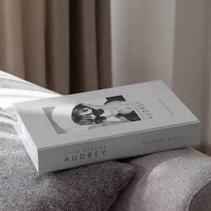 Könnte beinhalten: Ein Hardcover-Buch mit dem Titel "Little Book of Audrey" von Savanna Blake. Das Buch zeigt ein Schwarz-Weiß-Bild einer Frau auf dem Cover. Das Buch liegt auf einer grauen, gepolsterten Oberfläche.