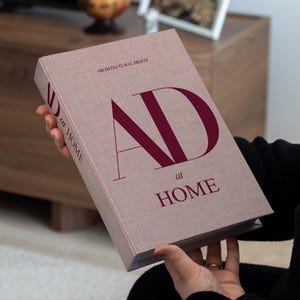 Può includere: Un libro con copertina rigida intitolato "AD at HOME" è tenuto in mano. La copertina è rosa tenue con grandi lettere bordeaux e le parole "ARCHITECTURAL DIGEST" in alto. Il dorso del libro è visibile e lo sfondo è sfocato.