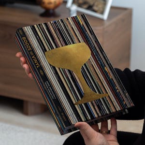 Può includere: Un libro intitolato "Booze & Vinyl" con un design di bicchiere da cocktail dorato. La copertina è realizzata con dischi in vinile vintage. Il libro è tenuto davanti a uno sfondo sfocato.