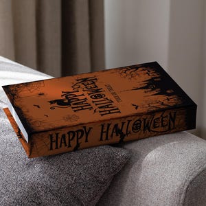 Puede incluir: Una caja rectangular con temática de Halloween con fondo naranja y texto negro que dice "Happy Halloween". La caja presenta un diseño espeluznante con un gato negro, murciélagos y una silueta de casa embrujada.