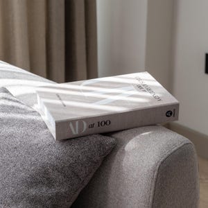 以下が含まれることがあります： 「AD at 100 A Century of Style」というタイトルのハードカバー本が、灰色の布張りのソファの上に置かれています。本は、白い文字とグラフィックデザインが施された、明るいベージュ色の表紙です。灰色のテクスチャード加工されたクッションが前景に見えます。