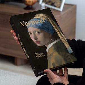 Könnte beinhalten: Ein gebundenes Buch mit dem Titel "Vermeer: The Complete Paintings" wird vor einem verschwommenen Hintergrund gehalten. Das Buchcover zeigt ein Porträt einer Frau mit blauem Kopftuch. Der Titel "Vermeer" ist in Goldbuchstaben gehalten.