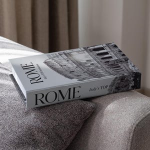 Può includere: Un libro intitolato "ROMA" con un'immagine in bianco e nero del Colosseo. Il libro è appoggiato su un divano grigio. È visibile anche il testo "Italy's TOP Destinations".