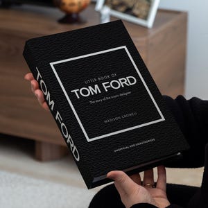Può includere: Un libro nero intitolato "Little Book of Tom Ford" con testo bianco e un bordo bianco. La copertina del libro ha un aspetto strutturato. Il libro è tenuto davanti a uno sfondo sfocato.