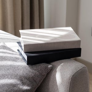 Puede incluir: Dos álbumes de fotos apilados uno encima del otro. El álbum superior es de color beige claro con una cubierta de tela texturizada. El álbum inferior es negro con una cubierta de tela lisa.