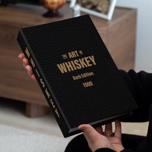 May include: A black book with the title "The Art in Whiskey" in gold lettering. The book is the "Dark Edition" from 1900. The book has a textured cover and is held by a person.