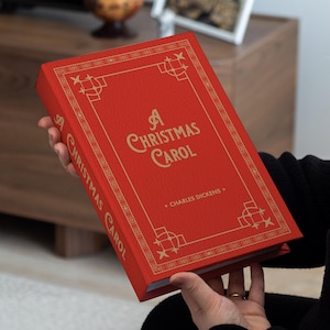 Può includere: Un libro rosso intitolato "A Christmas Carol" di Charles Dickens. Il libro ha lettere dorate e bordi decorativi. Il libro è tenuto in mano.