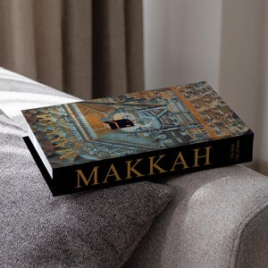 Può includere: Un libro con copertina rigida intitolato "MAKKAH" con un'immagine dettagliata di una città sulla copertina. Il libro è nero con scritte dorate e poggia su un cuscino grigio. La copertina presenta un'illustrazione architettonica dettagliata.